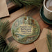 A decorative ornament featuring an illustration of a forest scene with an owl, placed on a wooden surface alongside a cup of coffee and some pine branches.