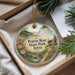 A decorative ornament with a scenic landscape of prairie roses, a small cabin, and a stream, hanging from a string.