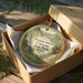 An oval ornament in a brown box, featuring a scenic landscape with a waterfall and trees, with the text "Preparation Canyon State Park, Iowa" printed on it.