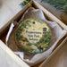 A decorative ornament in a box, featuring a painting of a scenic landscape with a small boat on a river, surrounded by trees and a tower.