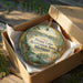 A wooden ornament in a box, featuring a scenic landscape with a lake and trees, with the text "Proud Lake Recreation Area Michigan" printed on it.
