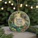 A round ornament with a painting of a scenic landscape, featuring a waterfall, a bird, and flowers, hangs in front of a Christmas tree with lights.