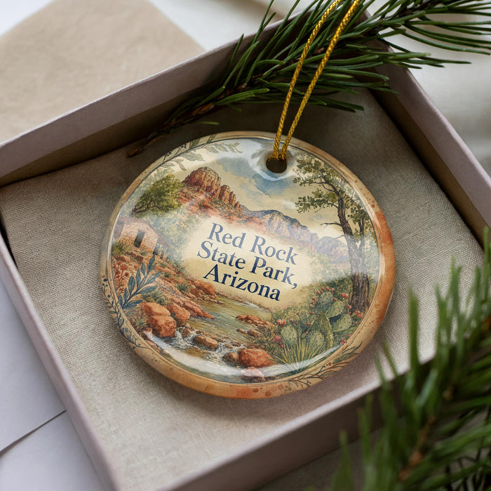 A decorative ornament with a landscape scene featuring a river, mountains, and desert vegetation, placed in a box with pine branches.