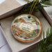 A decorative ornament with a landscape scene featuring a river, mountains, and desert vegetation, placed in a box with pine branches.