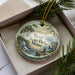 A decorative ornament in a box, featuring a scenic landscape with a waterfall and pine trees.
