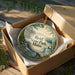 A decorative ornament in a brown box, featuring a scenic landscape with a waterfall and mountains, with the text "Reid State Park, Maine" printed on it.