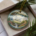 A decorative ornament with a scenic landscape of Rice Lake State Park in Minnesota, featuring a lake, mountains, and a cabin.
