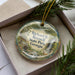 A decorative ornament in a box, featuring a scenic landscape with a waterfall, pine trees, and a cabin.