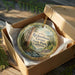 An oval ornament in a brown box, featuring a scenic image of a forest and a stream, with the text "Richardson Grove State Park, California" printed on it.