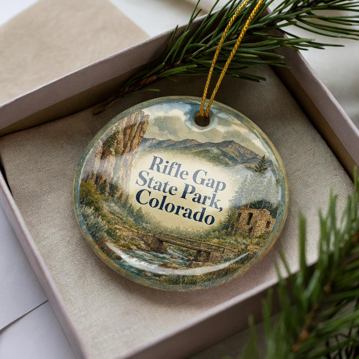 A decorative ornament in a box, featuring a scenic landscape with mountains, a river, and pine trees.