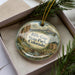 A decorative ornament in a box, featuring a scenic landscape with mountains, a river, and pine trees.