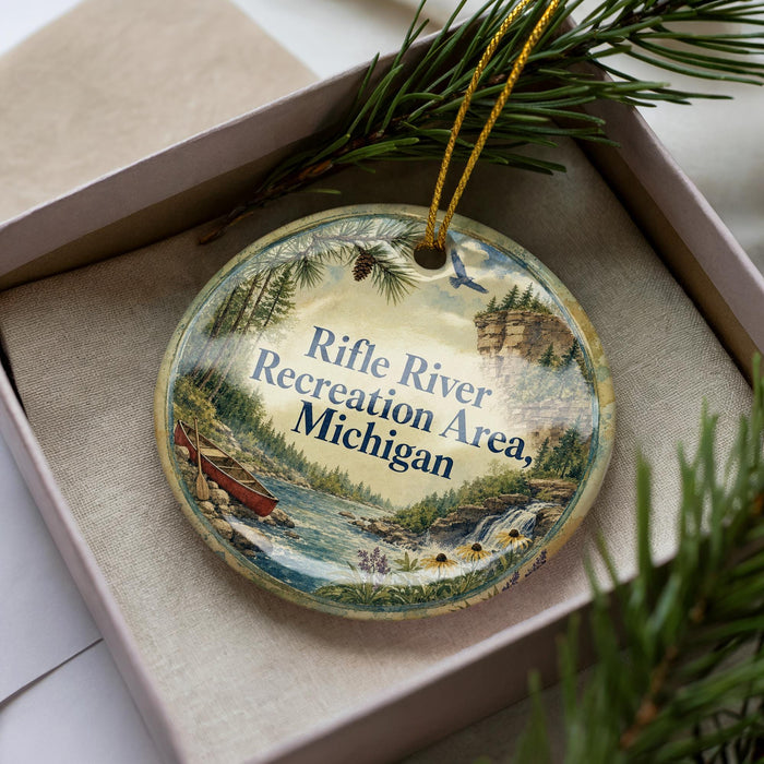 A decorative ornament with a scenic image of a river and forest, featuring a rifle and a boat, is displayed in a box with pine branches.