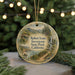 A round ornament with a painting of a scenic landscape, featuring a cabin and a waterfall, hangs in front of a blurred background of Christmas lights.