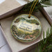 A decorative ornament in a box, featuring a scenic landscape with a river, trees, and a lighthouse, with the text "Robinson State Park, Massachusetts" printed on it.