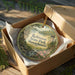 An ornament in a box, featuring a scenic landscape with a river, trees, and a building, likely representing Robinson State Park in Massachusetts.
