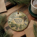 A round ornament with a scenic illustration of Roland Cooper State Park, Alabama, is displayed on a wooden surface alongside a cup of coffee and some pine branches.