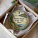 A round ornament in a box, featuring a scenic landscape with a path leading to a cliff overlooking the water.
