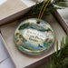 A decorative ornament with a landscape scene featuring a lake, mountains, and pine trees, along with a bird in the water.