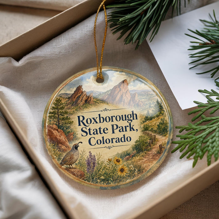 A decorative ornament with a scenic landscape of rocky mountains, pine trees, and wildflowers, featuring the text "Roxborough State Park, Colorado" on it.
