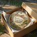 A decorative ornament in a box, featuring a scenic landscape of mountains and a bird, with the text "Roxborough State Park, Colorado" printed on it.