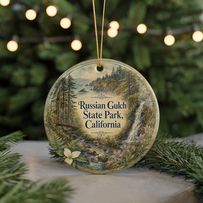 A round ornament with a painting of a scenic landscape, featuring a waterfall, pine trees, and a bridge, hangs in front of a Christmas tree with twinkling lights.
