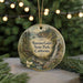 A round ornament with a painting of a scenic landscape, featuring a waterfall, pine trees, and a bridge, hangs in front of a Christmas tree with twinkling lights.