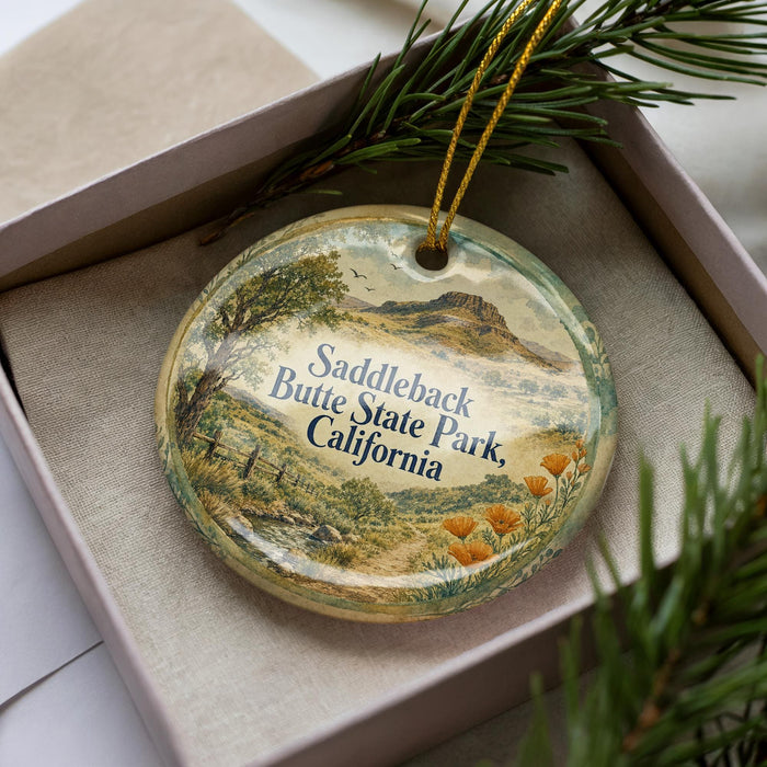 A decorative ornament in a box, featuring a scenic landscape with a mountain and trees, along with the text "Saddleback Butte State Park, California".