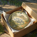 A round ornament in a brown box, featuring a scenic landscape with a mountain and trees, is displayed on a wooden surface.