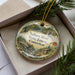 A decorative ornament in a box, featuring a scenic landscape with a river, forest, and bridge.