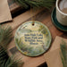 A decorative ornament with a lake scene and birds, placed on a wooden surface next to a cup of coffee and some pine branches.