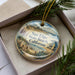 A decorative ornament in a box, featuring a beach scene with palm trees, a lifeguard tower, and a sandy beach.