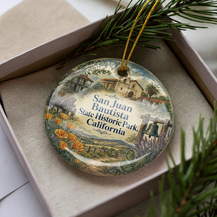 A decorative ornament with a scenic illustration of Santa Juan Bautista State Historic Park, California, displayed in a box with pine branches.