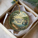 A decorative ornament in a box, featuring a scenic landscape with a lake, trees, and a small building.