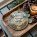 A decorative ornament featuring a lighthouse and a coastal scene, placed on a wooden tray alongside pine cones and a lit candle.