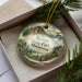 A decorative ornament with a scenic landscape of mountains, trees, and a winding path, placed inside a box with pine branches.