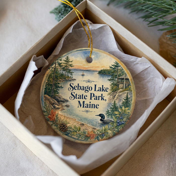 A decorative ornament in a box, featuring a scenic landscape with a lake, trees, and a bird, along with the text "Sebago Lake State Park, Maine".