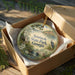 A decorative ornament in a box, featuring a scenic landscape with a lake, trees, and a bird, is displayed on a wooden surface.