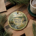 A decorative ornament with a scenic image of Seneca Creek State Park, Maryland, featuring a bird, a bridge, and flowers, is displayed on a wooden surface alongside a cup of coffee and some pine branches.