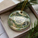 A decorative ornament in a box, featuring a scenic landscape with a river, trees, and a house, with the text "Shakamak State Park, Indiana" printed on it.