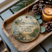A round ornament with a lighthouse and a seagull, surrounded by pine cones and a lit candle, sits on a wooden tray.