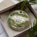 A decorative ornament in a box, featuring a scenic landscape with a river, trees, and mountains.