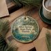 A round ornament with a painting of Silver Springs State Park, Florida, featuring a manatee and a bird, is displayed on a wooden surface alongside a cup of coffee and some pine branches.
