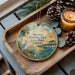 A decorative ornament featuring a seagull, palm trees, and a pier, with the text "Skyway Fishing Pier State Park, Florida" displayed on it. The ornament is placed on a wooden tray, with pine cones and a lit candle nearby.