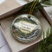 A decorative ornament in a box, featuring a scenic landscape with mountains, waterfalls, and greenery.