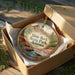 A wooden ornament in a box, featuring a scenic landscape with mountains, water, and greenery.
