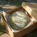 A decorative ornament in a box, featuring a scenic landscape with a lake, trees, and a bird, is placed on a wooden surface.