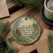 A round ornament with a forest scene and text, placed on a wooden surface next to a cup of coffee and some pine branches.