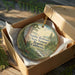 A round ornament in a brown box, featuring a scenic landscape with trees and a river, with the text "Smiththe Redwoods State Natural Reserve, California" printed on it.