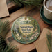 A decorative ornament featuring a scenic image of Smithall Woods State Park in Georgia, with a deer and flowers, is displayed on a wooden surface alongside a cup of coffee and some pine branches.