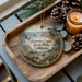 A decorative ornament featuring a scenic coastal landscape with a lighthouse, pine cones, and a lit candle on a wooden tray.